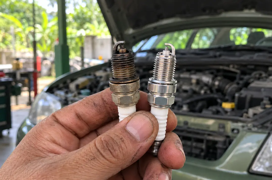 A mechanic's hand comparing two spark plugs by an open engine